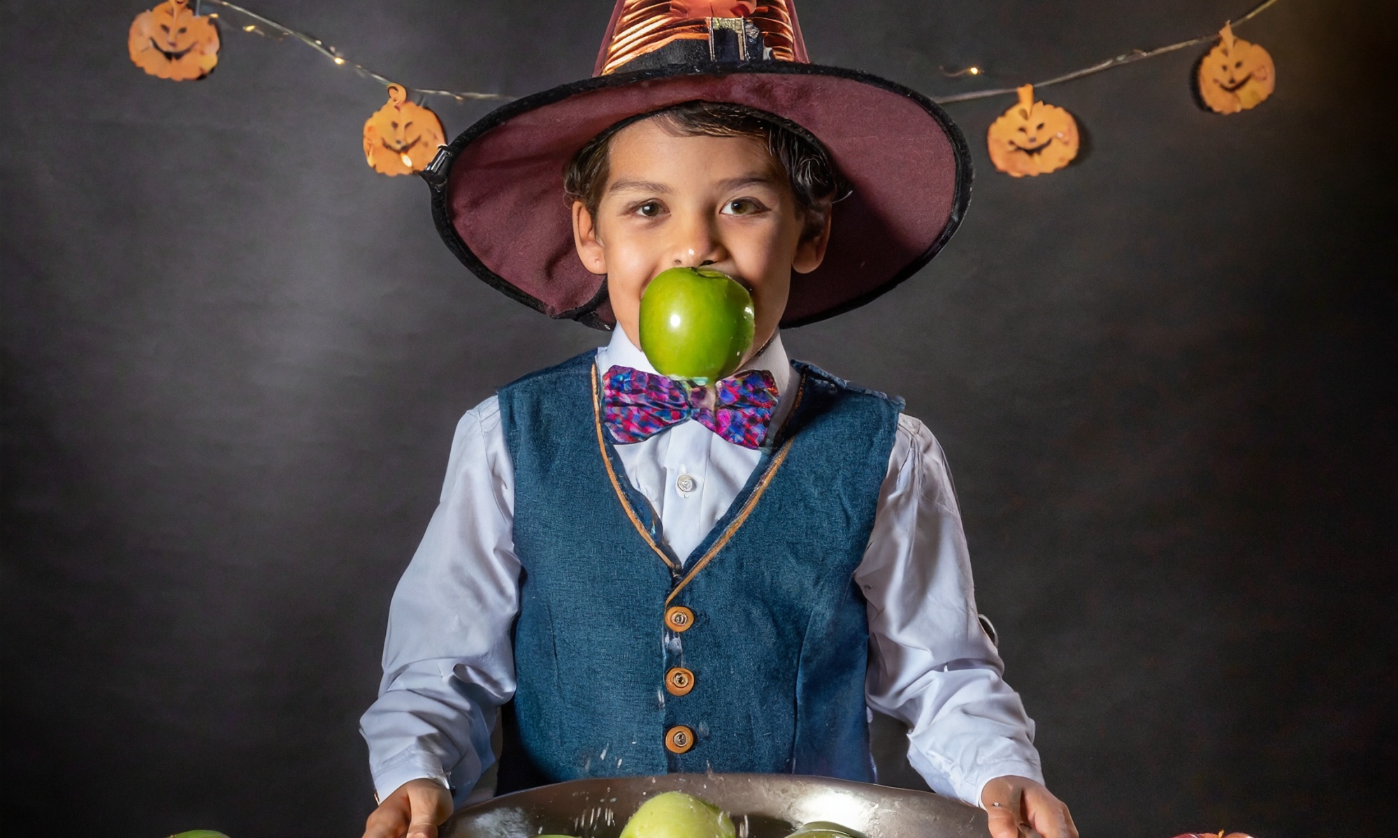 Kind im Halloweenkostüm hat einen Apfel im Mund vor einer großen Schüssel mit Wasser
